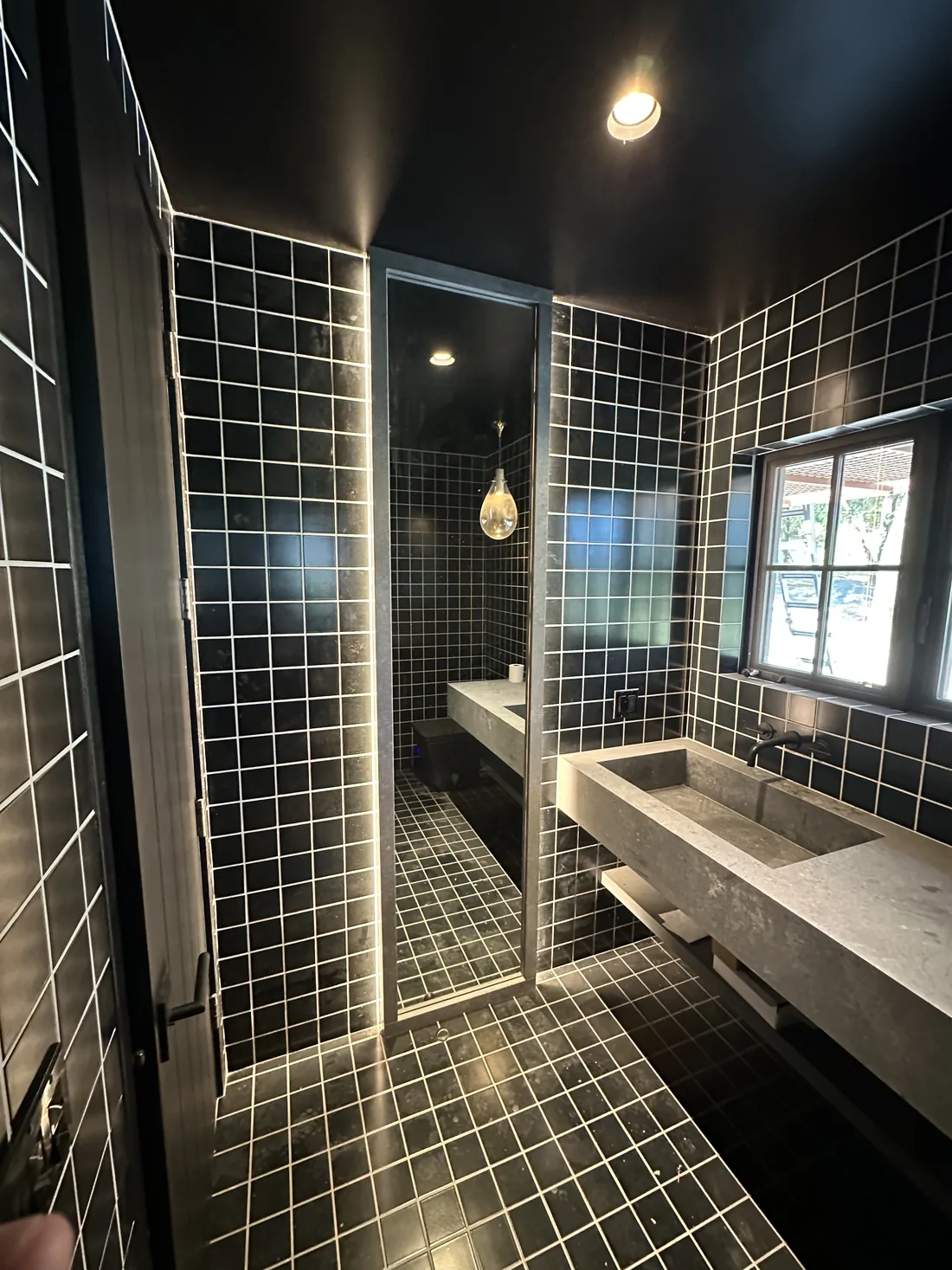 Narrow black-tiled bathroom with a concrete trough sink, a ceiling wall-pack fixture above and a recessed downlight plus a glass-shade pendant in the shower beyond, in Sonoma County